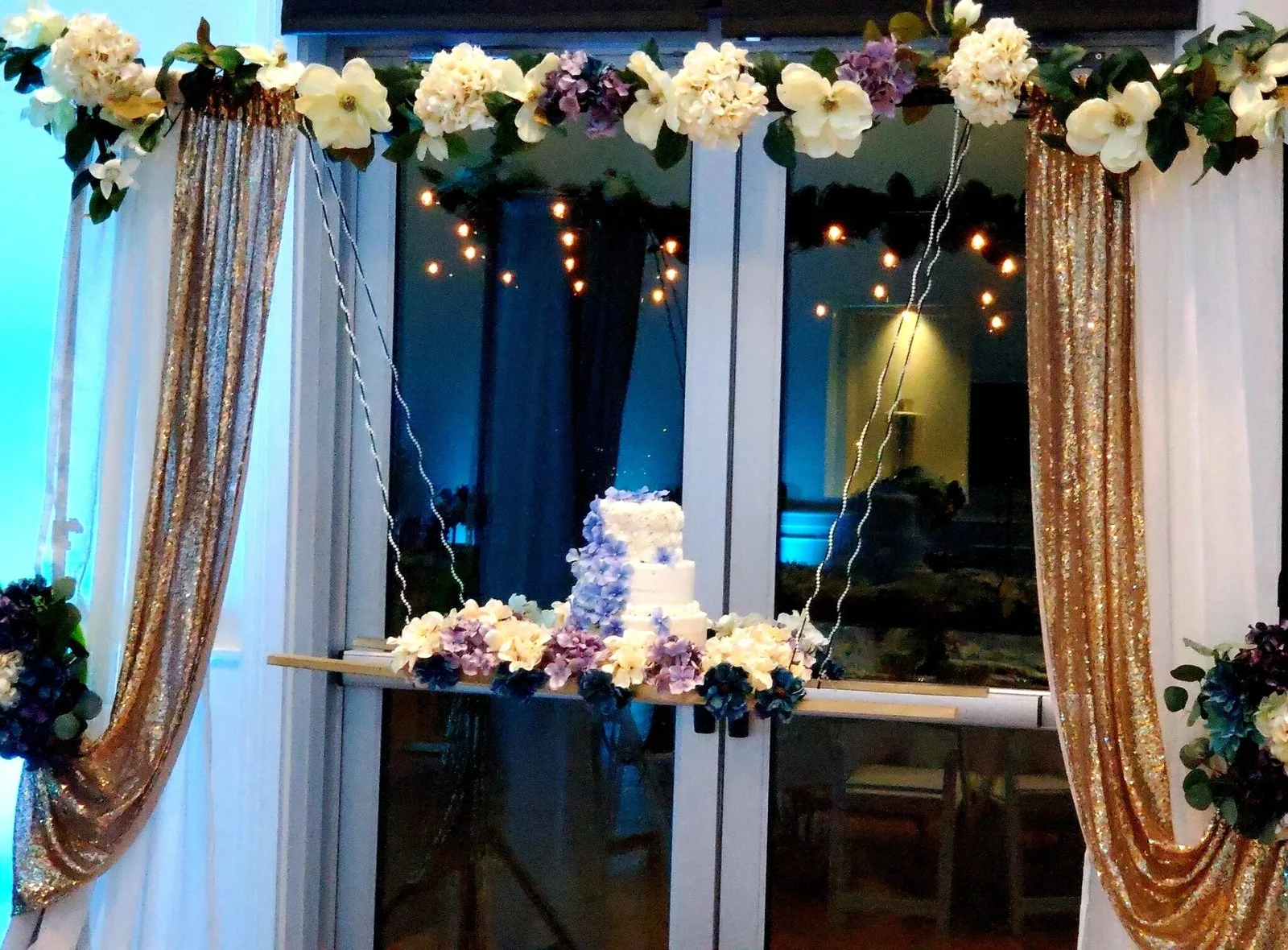 A birthday cake table framed by white flowers and gold draping